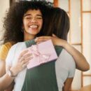 An excited young woman with curly hair hugging her friend and smiling after receiving a gift in a pink box.