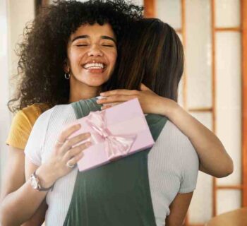 An excited young woman with curly hair hugging her friend and smiling after receiving a gift in a pink box.