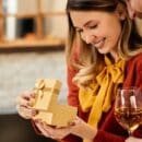 A happy woman opens a gift box from her smiling boyfriend as he stands next to her, holding a glass of white wine.