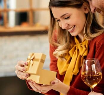 A happy woman opens a gift box from her smiling boyfriend as he stands next to her, holding a glass of white wine.