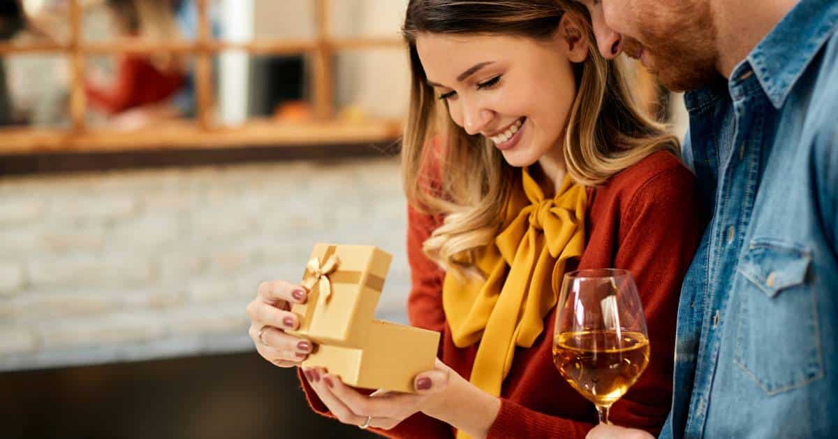A happy woman opens a gift box from her smiling boyfriend as he stands next to her, holding a glass of white wine.