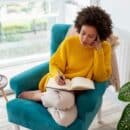 A woman relaxes in an armchair in her living room while holding a pen and thinking about what to write in her journal.
