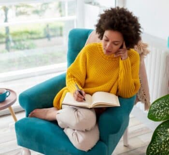 A woman relaxes in an armchair in her living room while holding a pen and thinking about what to write in her journal.