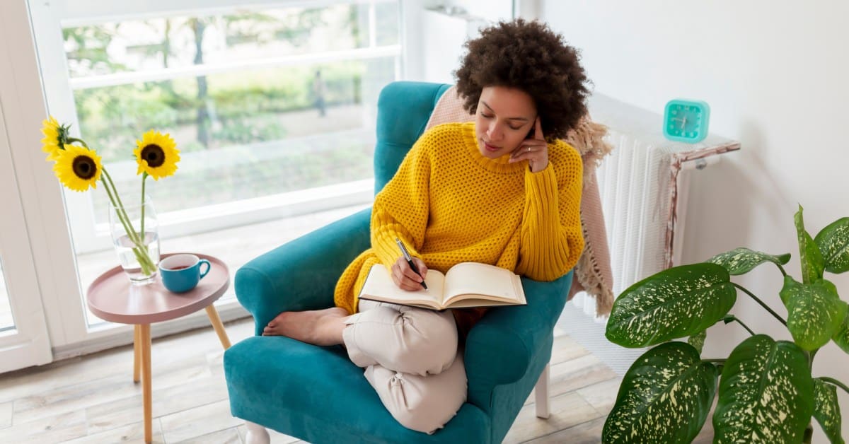 A woman relaxes in an armchair in her living room while holding a pen and thinking about what to write in her journal.