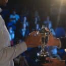 A backstage view of a smiling business man presenting a trophy to another business man as a crowd watches in the distance.