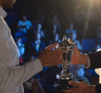 A backstage view of a smiling business man presenting a trophy to another business man as a crowd watches in the distance.