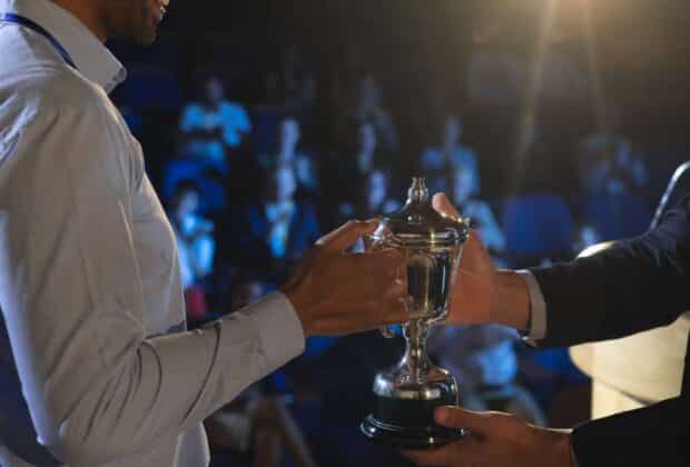 A backstage view of a smiling business man presenting a trophy to another business man as a crowd watches in the distance.