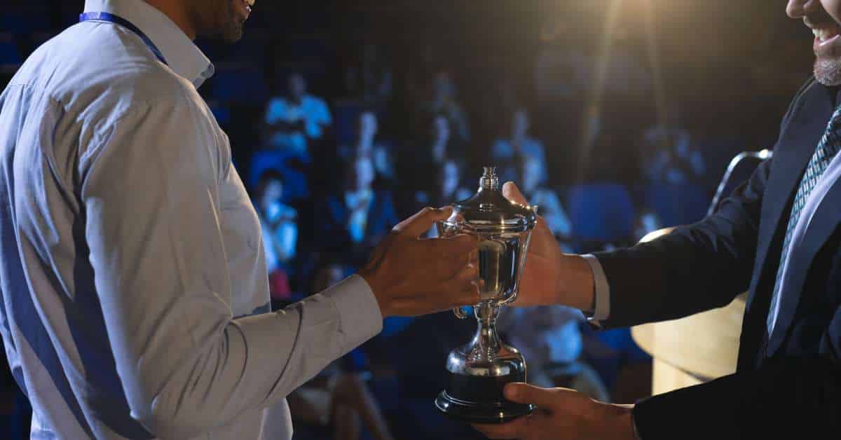 A backstage view of a smiling business man presenting a trophy to another business man as a crowd watches in the distance.