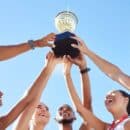 A group of young male and female athletes smiling. They each place a hand on a trophy and hold it up in the air.