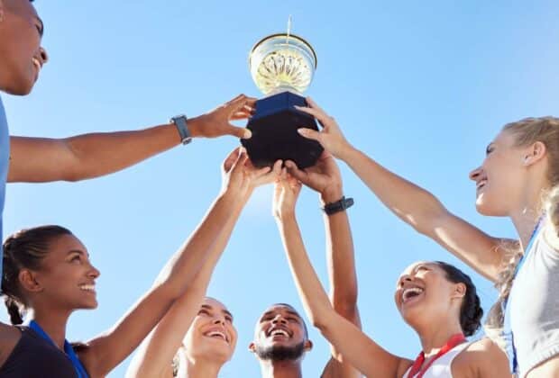 A group of young male and female athletes smiling. They each place a hand on a trophy and hold it up in the air.