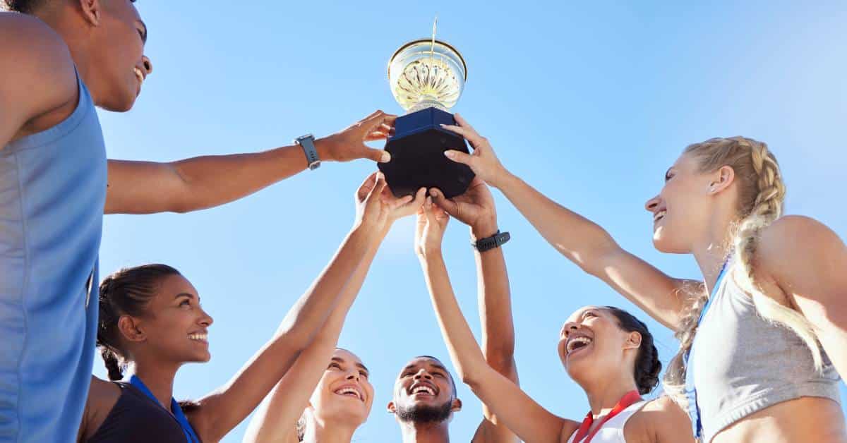 A group of young male and female athletes smiling. They each place a hand on a trophy and hold it up in the air.