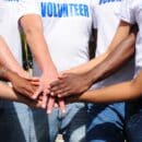 A group of seven people put their hands together. They all wear denim jeans and white t-shirts that read "Volunteer."