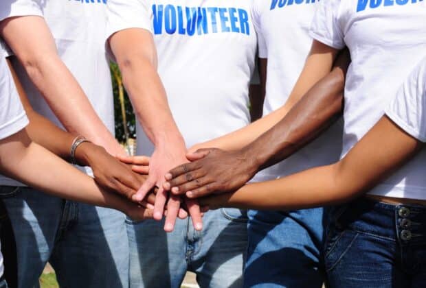 A group of seven people put their hands together. They all wear denim jeans and white t-shirts that read "Volunteer."