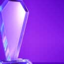 A blank crystal award sits on a solid surface against a purple gradient background. Light reflects off the award.