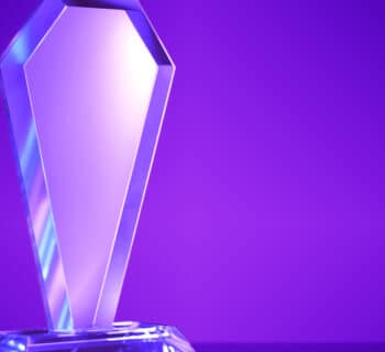 A blank crystal award sits on a solid surface against a purple gradient background. Light reflects off the award.