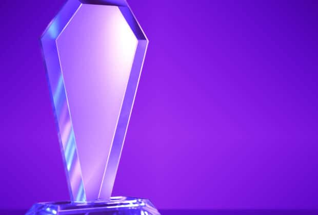 A blank crystal award sits on a solid surface against a purple gradient background. Light reflects off the award.