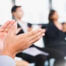 A group of blurred business professionals sitting in a circle, clapping. Bright sunlight shines through a wall of windows.