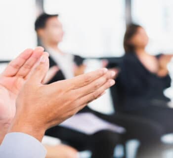 A group of blurred business professionals sitting in a circle, clapping. Bright sunlight shines through a wall of windows.