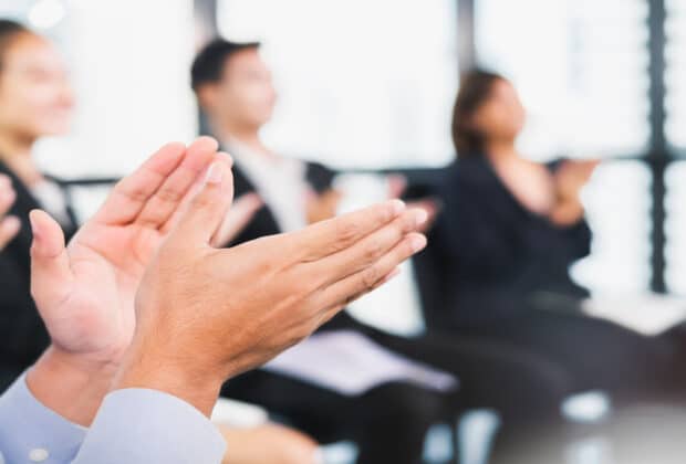 A group of blurred business professionals sitting in a circle, clapping. Bright sunlight shines through a wall of windows.
