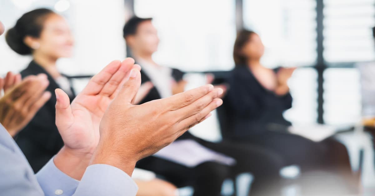 A group of blurred business professionals sitting in a circle, clapping. Bright sunlight shines through a wall of windows.
