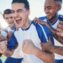 A group of five young men wearing soccer jerseys stand close together and cheer in celebration near the soccer goal.