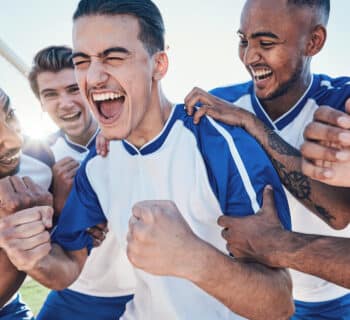 A group of five young men wearing soccer jerseys stand close together and cheer in celebration near the soccer goal.