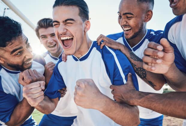 A group of five young men wearing soccer jerseys stand close together and cheer in celebration near the soccer goal.