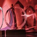 Four transparent awards of unique shapes with wood bases reflect shades of light and shadowing on a red background.