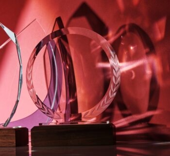 Four transparent awards of unique shapes with wood bases reflect shades of light and shadowing on a red background.