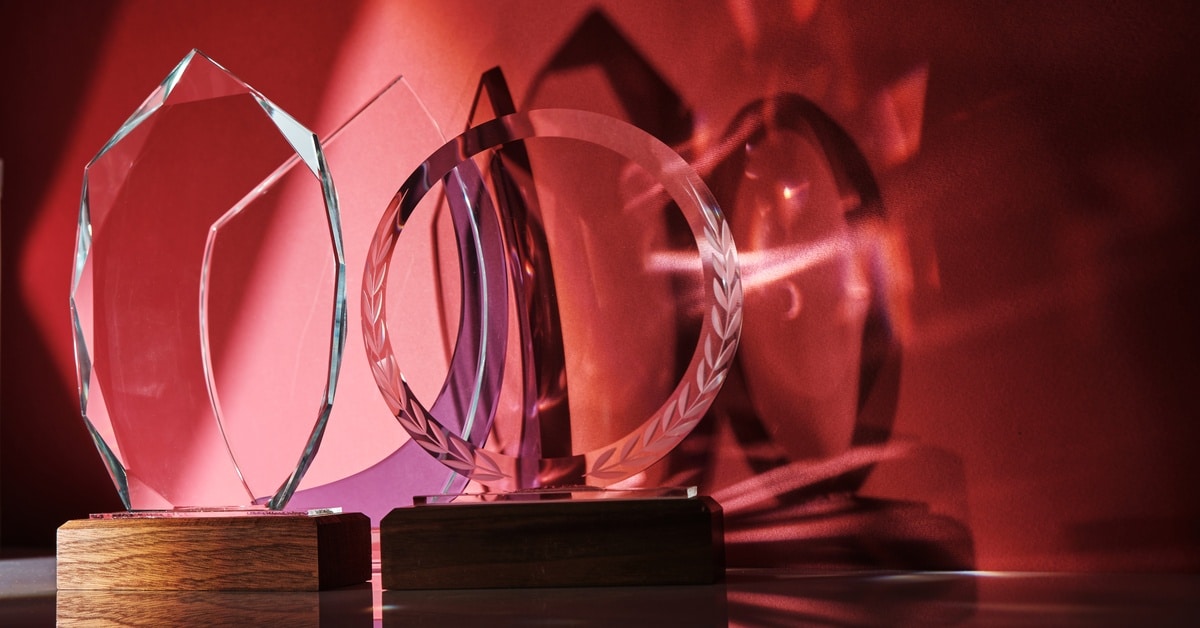 Four transparent awards of unique shapes with wood bases reflect shades of light and shadowing on a red background.