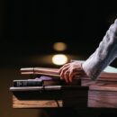 A close-up of a pastor standing with his hands on a pulpit in front of a dark room. A tablet and Bible lay on the stand.
