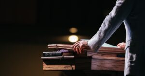 A close-up of a pastor standing with his hands on a pulpit in front of a dark room. A tablet and Bible lay on the stand.