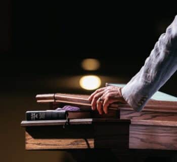 A close-up of a pastor standing with his hands on a pulpit in front of a dark room. A tablet and Bible lay on the stand.