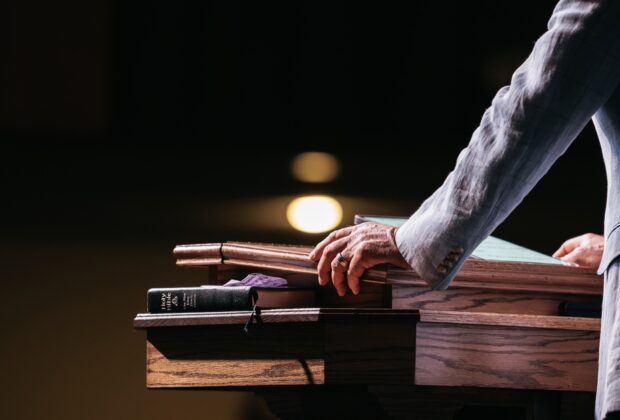 A close-up of a pastor standing with his hands on a pulpit in front of a dark room. A tablet and Bible lay on the stand.